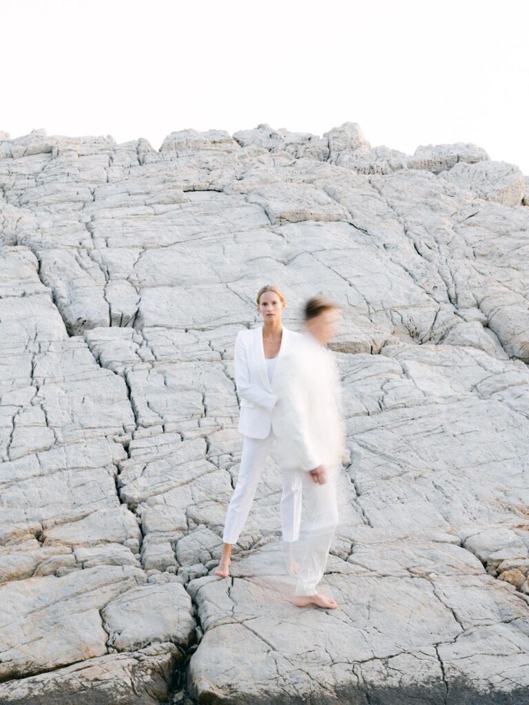 Stylish wedding couple on the French Riviera: the bride, wearing a sleek white blazer, gazes confidently at the camera, while the groom, also in white, walks past in a motion blur. Minimalist and modern composition with a bright, mineral backdrop.